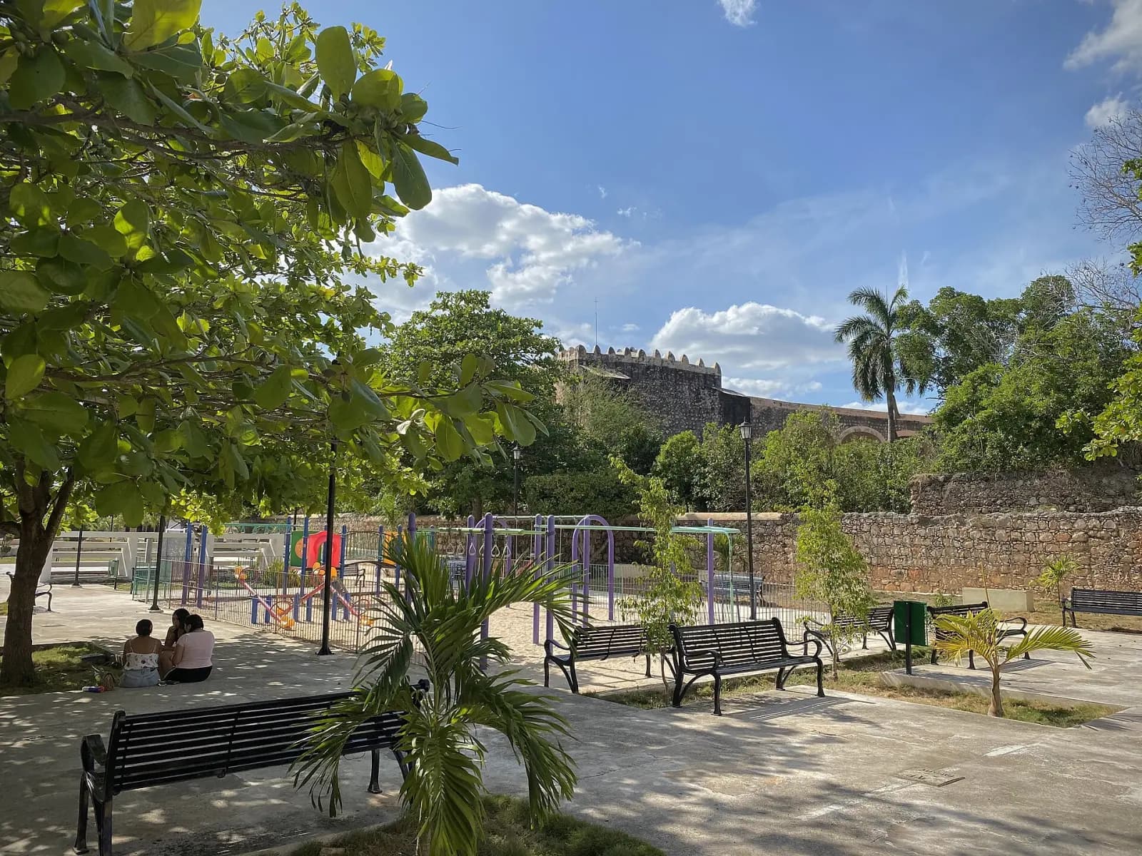 Parque de Maní con bancas y juegos infantiles; al fondo, los muros del exconvento