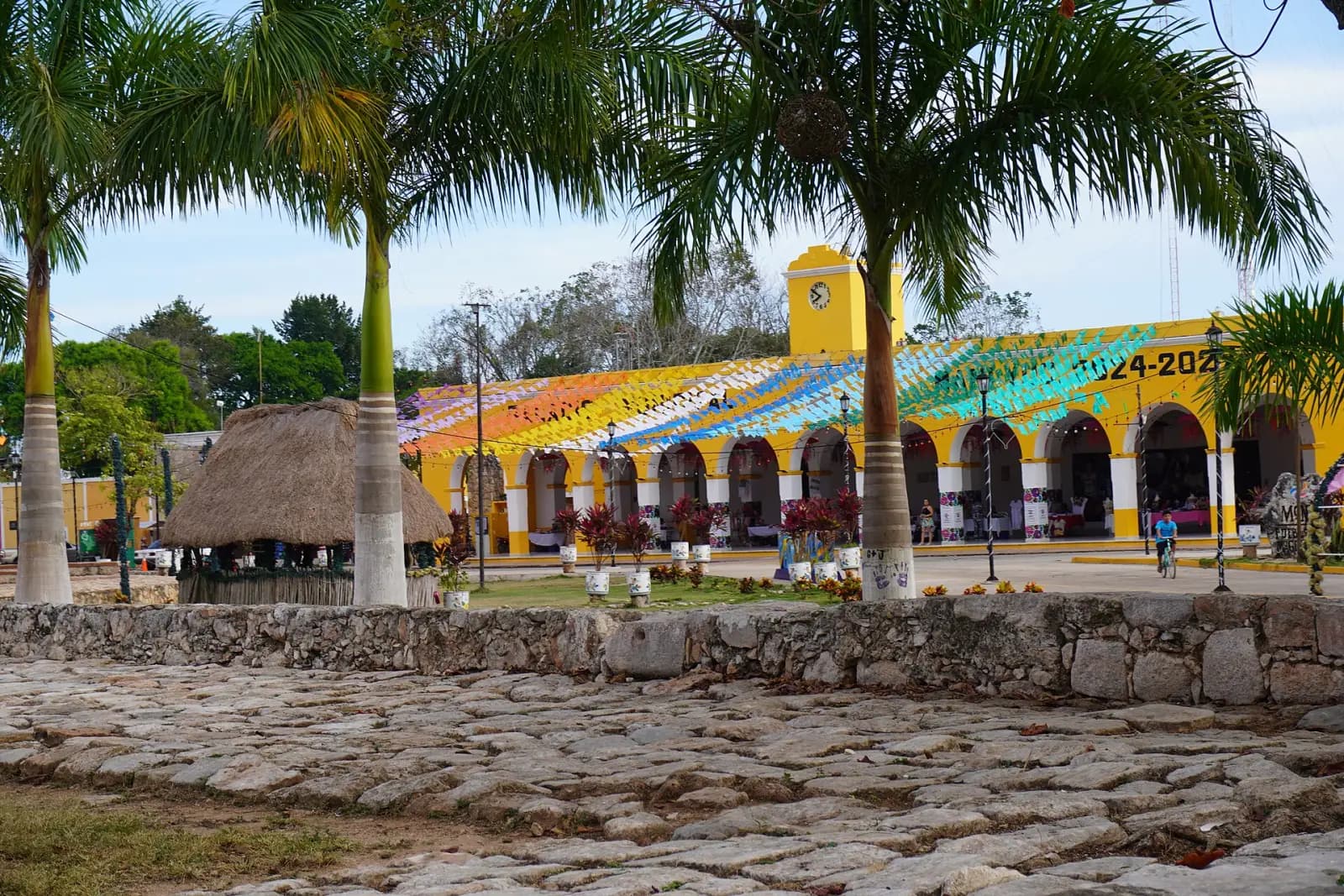 Portales del palacio municipal de Maní pintados de colores, con palmeras y piso de piedra en la plaza